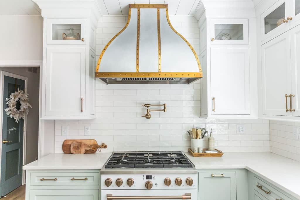 Nicholas Design Build | Modern kitchen interior featuring white cabinetry, subway tile backsplash, and a prominent gold-trimmed range hood over a gas stove.