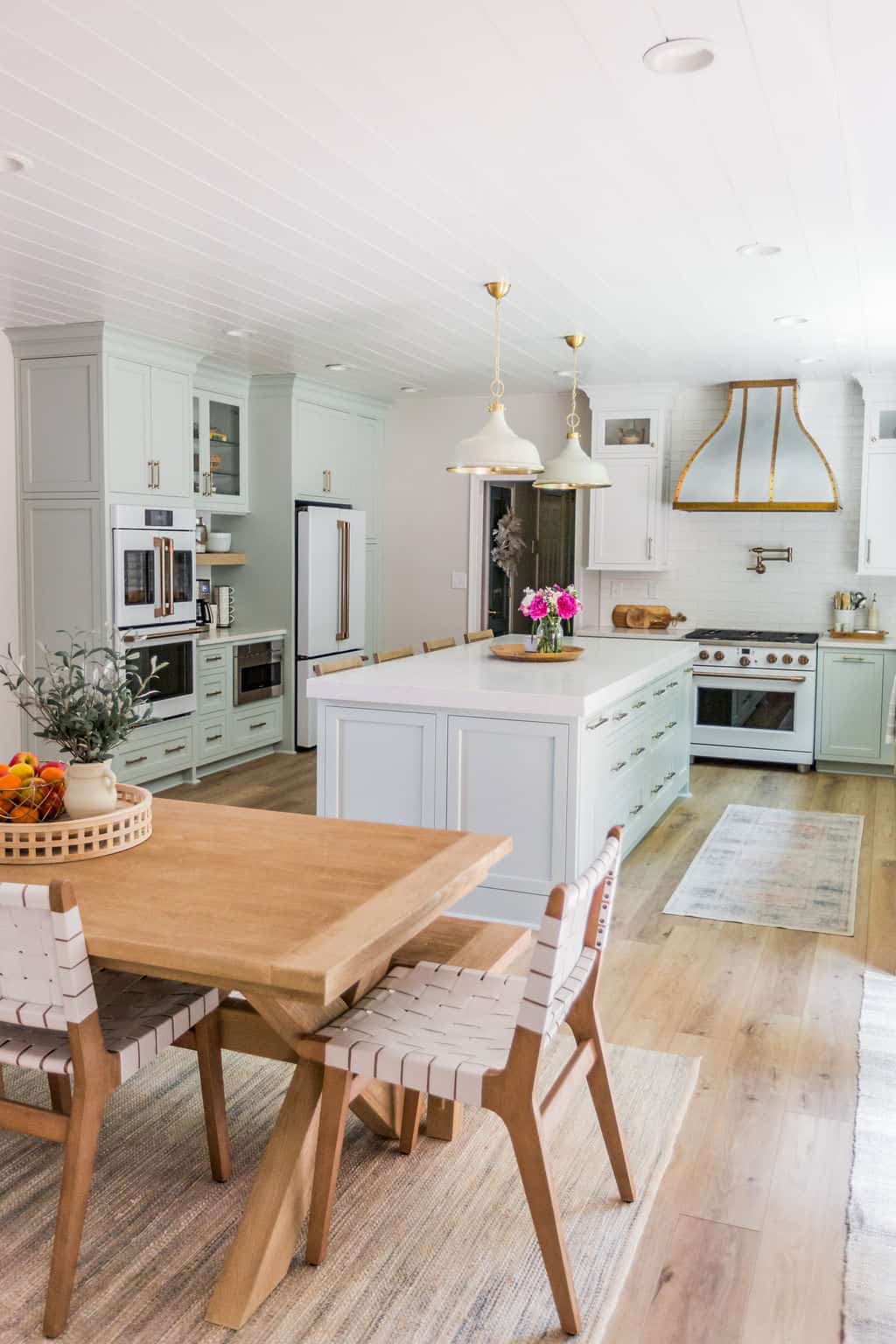 Nicholas Design Build | A bright and modern kitchen with white cabinetry, stainless steel appliances, and a central island, adorned with pendant lighting and a wooden dining table set.