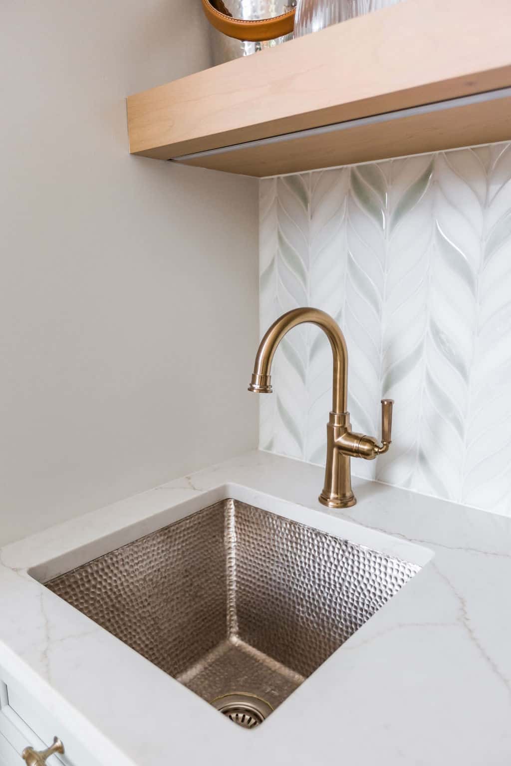 Nicholas Design Build | Modern kitchen sink with a hammered metal basin, white marble countertop, and a bronze faucet against a tiled backsplash with leaf pattern.