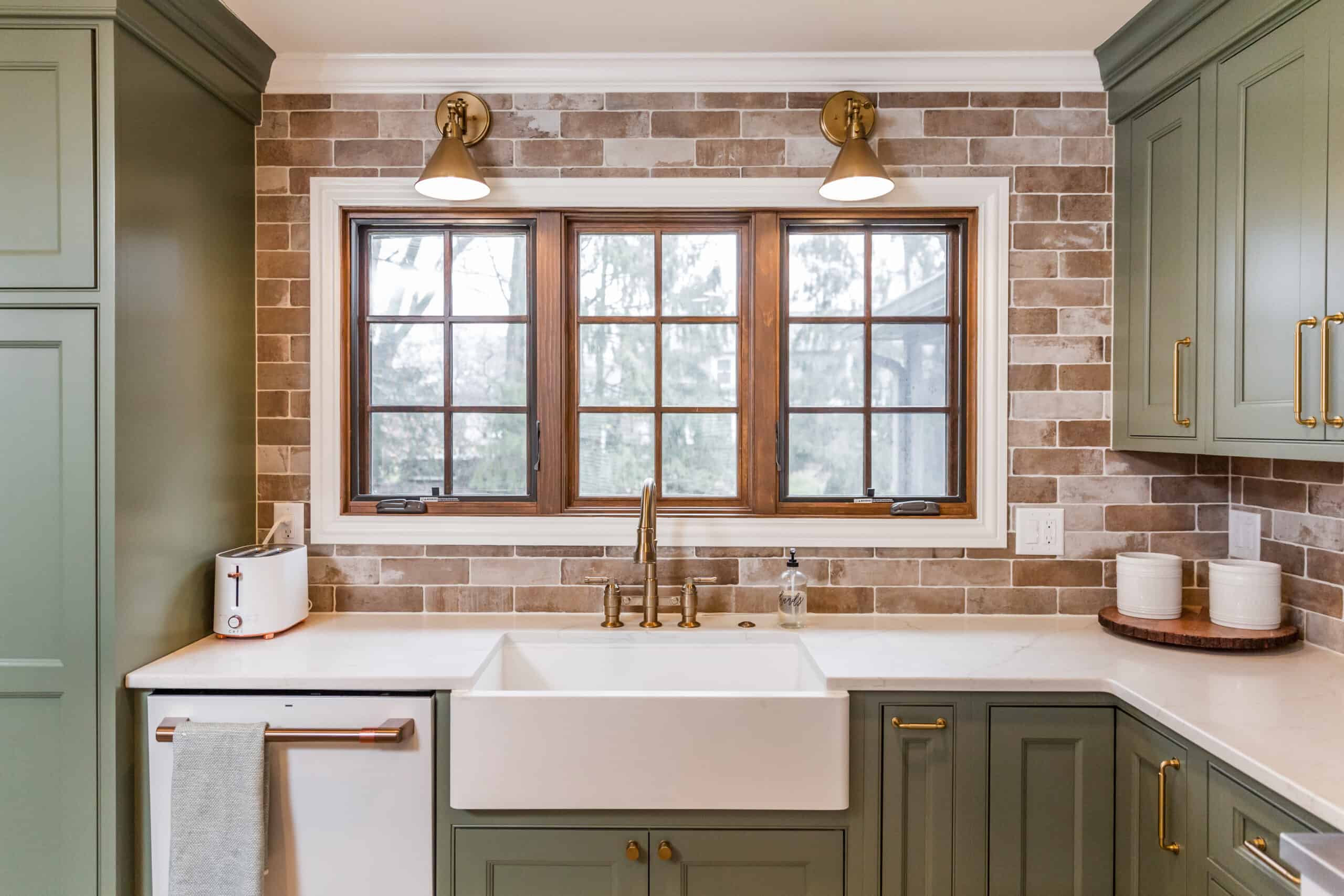 Nicholas Design Build | A modern kitchen with green cabinetry, white countertops, and a window framed by brick-patterned backsplash with two wall-mounted lights above the sink.