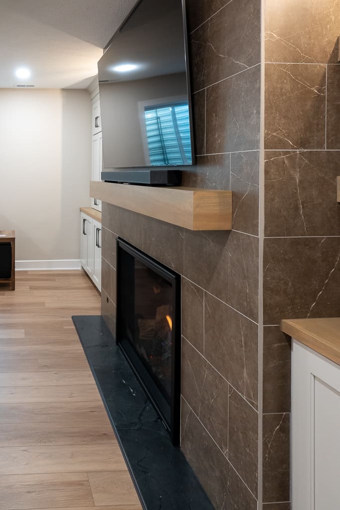 Nicholas Design Build | A modern fireplace with a mounted television above it, flanked by a wood mantle and beige tiles, in a room with hardwood flooring.