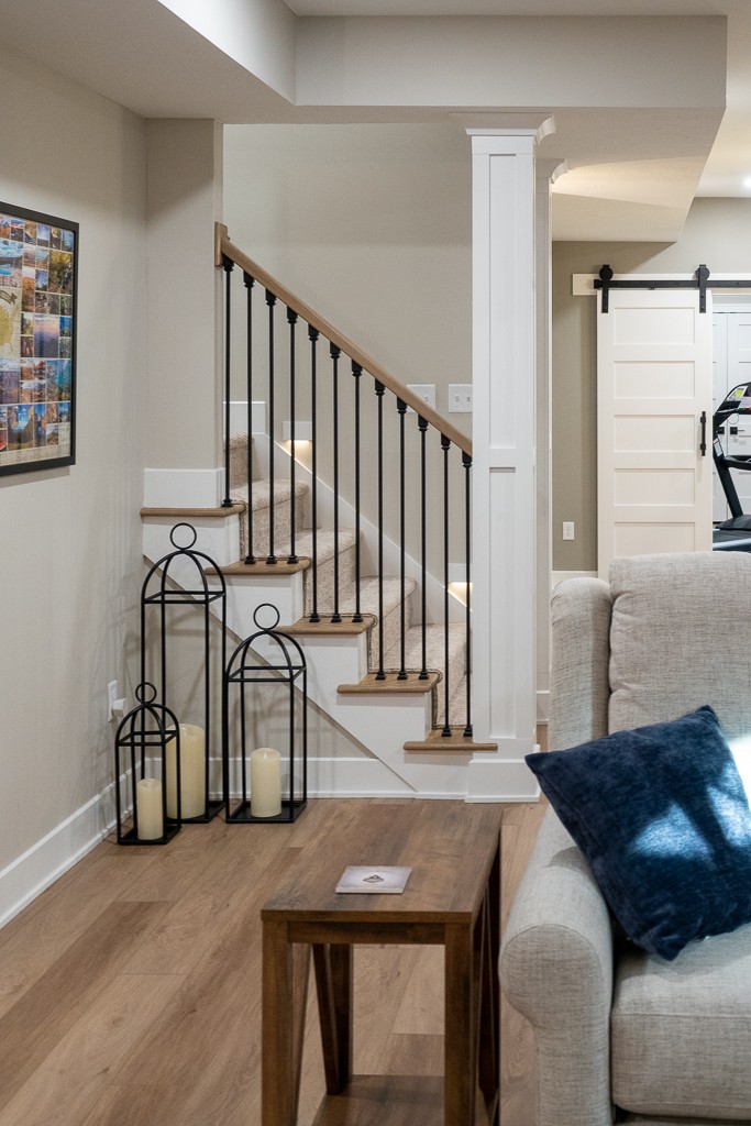 Nicholas Design Build | An elegant interior hallway featuring a wooden staircase with white balusters, a plush sofa with a blue pillow, a wooden coffee table, and decorative lanterns beside the stairs.