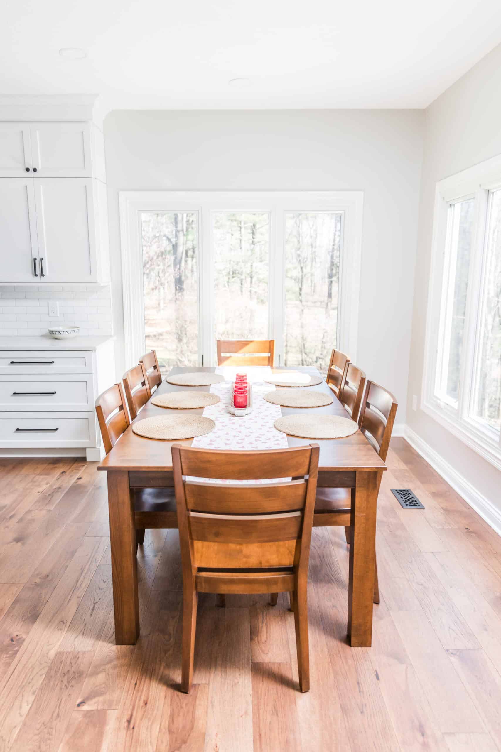 Nicholas Design Build | Brightly lit kitchen nook with a round wooden table and brown chairs, large windows showcasing a wooded view, and white cabinetry.