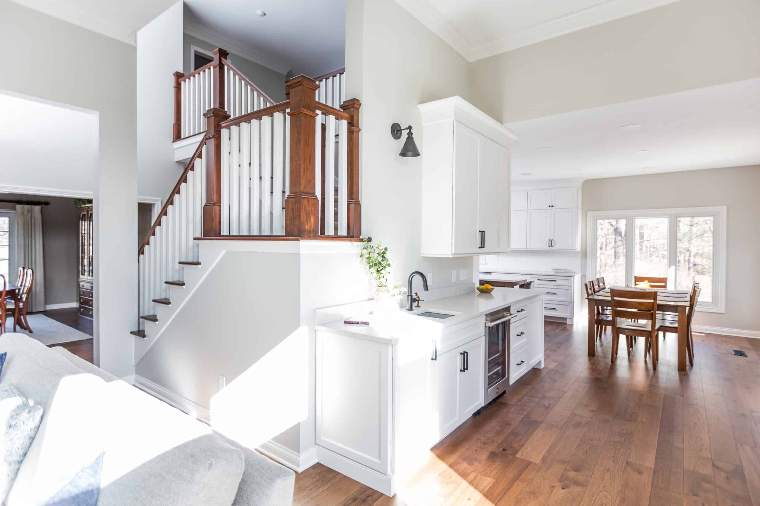 Nicholas Design Build | Modern kitchen with white cabinetry and wooden floors, featuring a staircase with wooden rails and a dining area in the background.