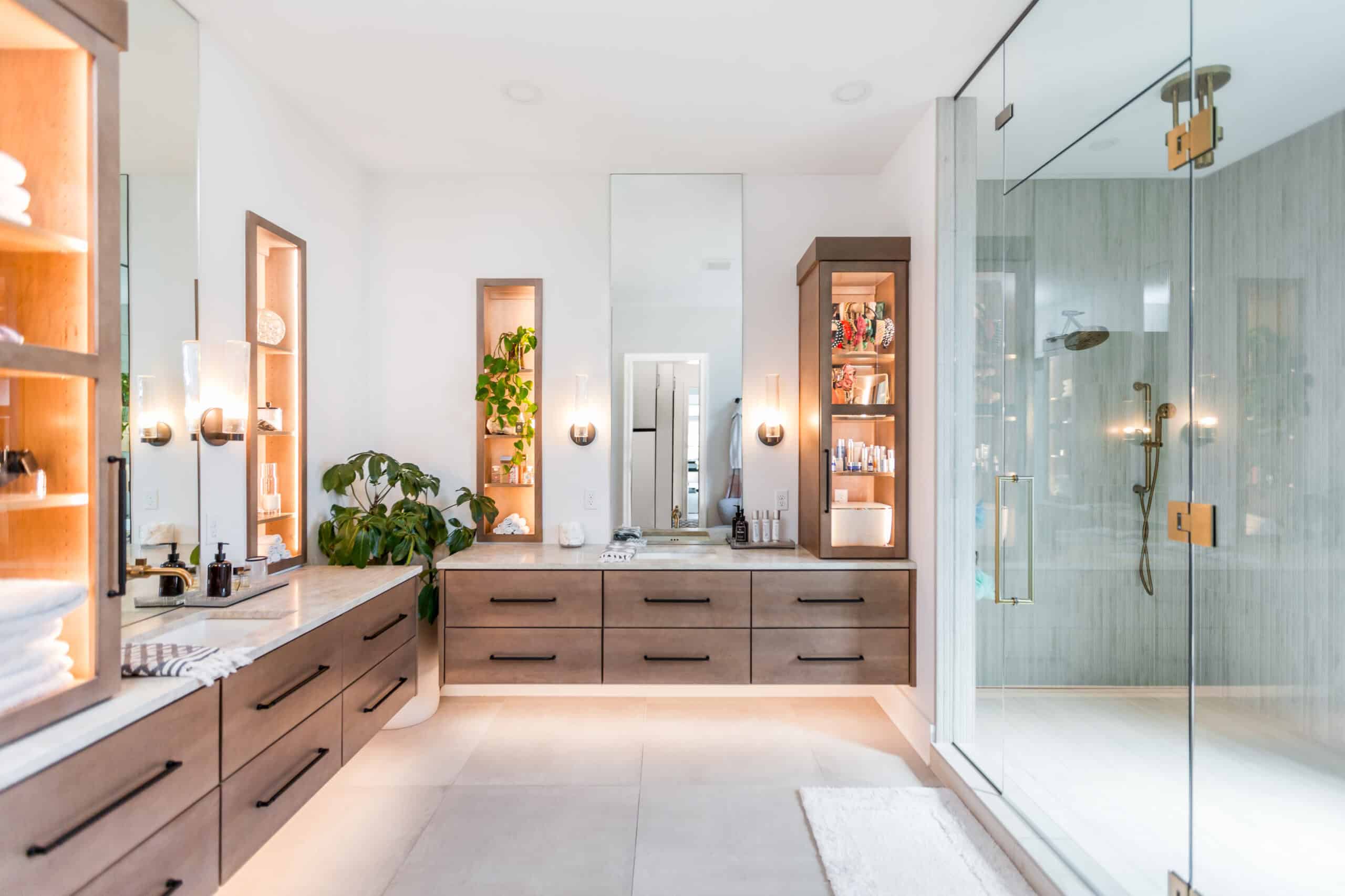 Nicholas Design Build | A modern bathroom with a double sink vanity, illuminated shelving, large mirror, potted plants, and a glass-enclosed shower.