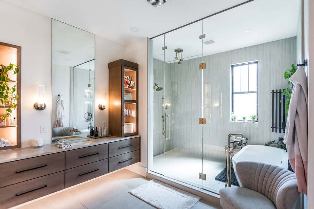 Nicholas Design Build | Modern bathroom with a glass-enclosed shower, freestanding bathtub, double vanity with large mirrors, wooden cabinets, potted plants, gray chair, and hanging bathrobes. Natural light from a window.