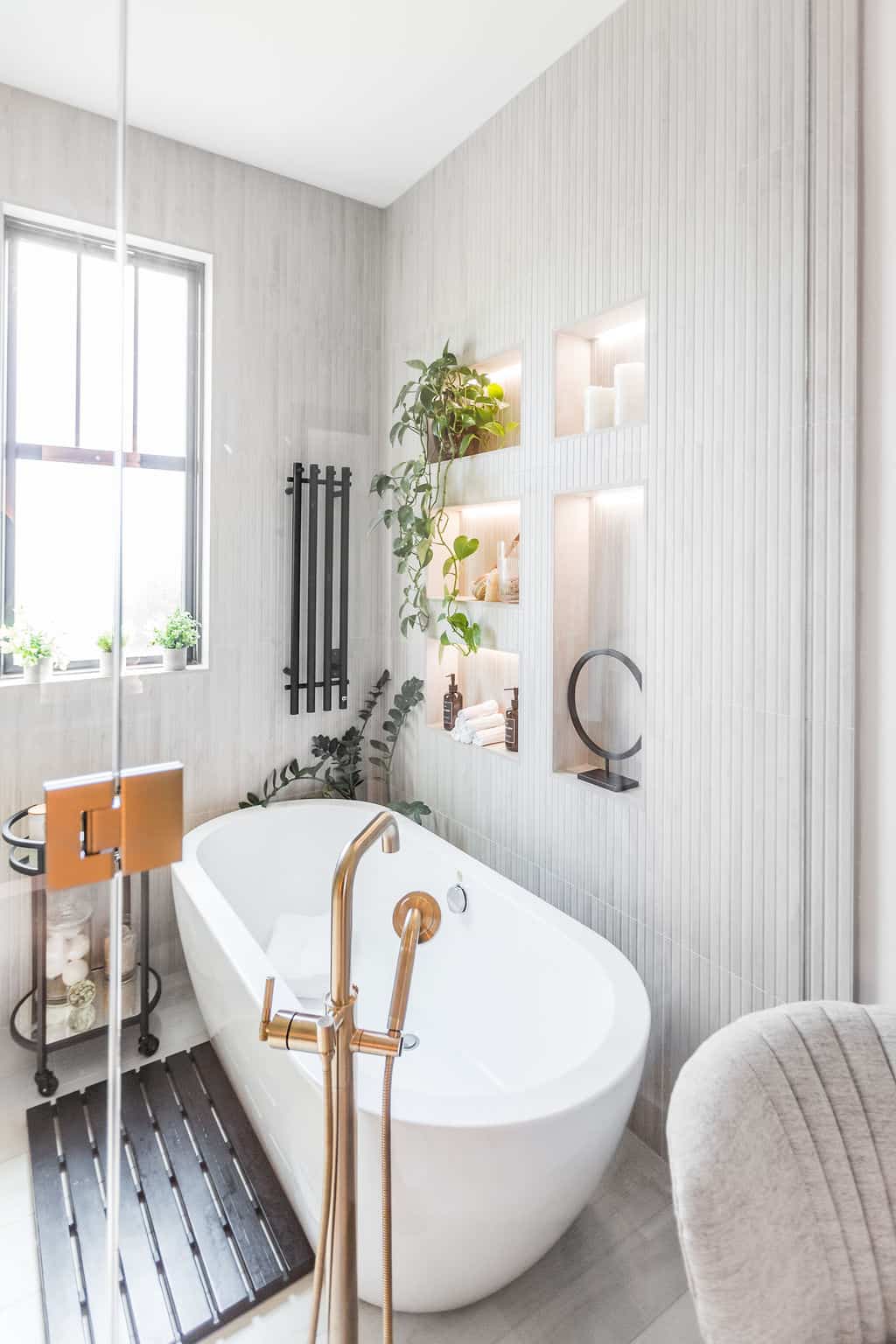 Nicholas Design Build | A modern bathroom with a white freestanding bathtub, gold fixtures, potted plants, a towel rack, wall niches with decor, glass shower wall, and a window allowing natural light.