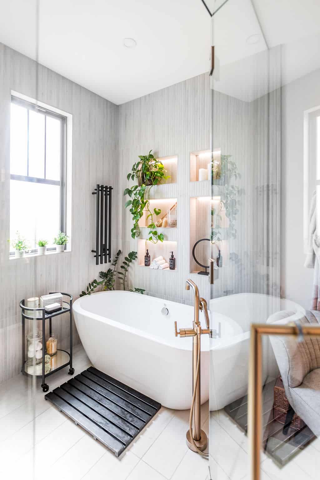 Nicholas Design Build | A modern bathroom with a white freestanding bathtub, wooden floors, plants, a black towel rack, and a window on the left. Shelves with illuminated plants and toiletries are on the wall behind the tub.