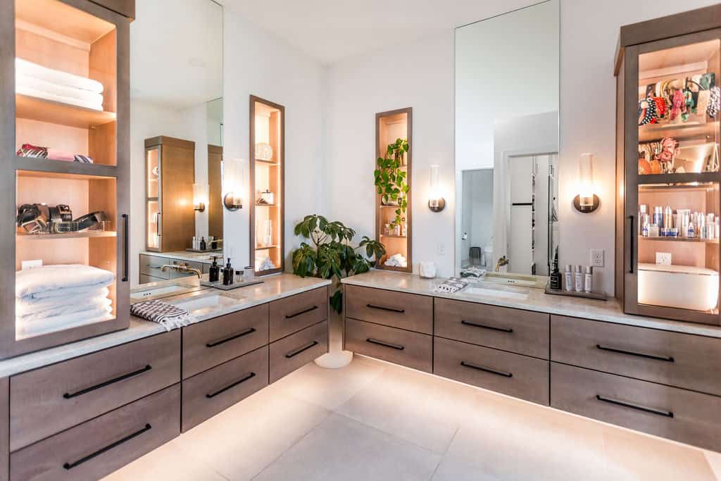 Nicholas Design Build | Modern bathroom with double sinks, large mirrors, wooden cabinetry, open shelving, and a potted plant. Towels, toiletries, and decor items are neatly arranged on the shelves. Neutral color palette.