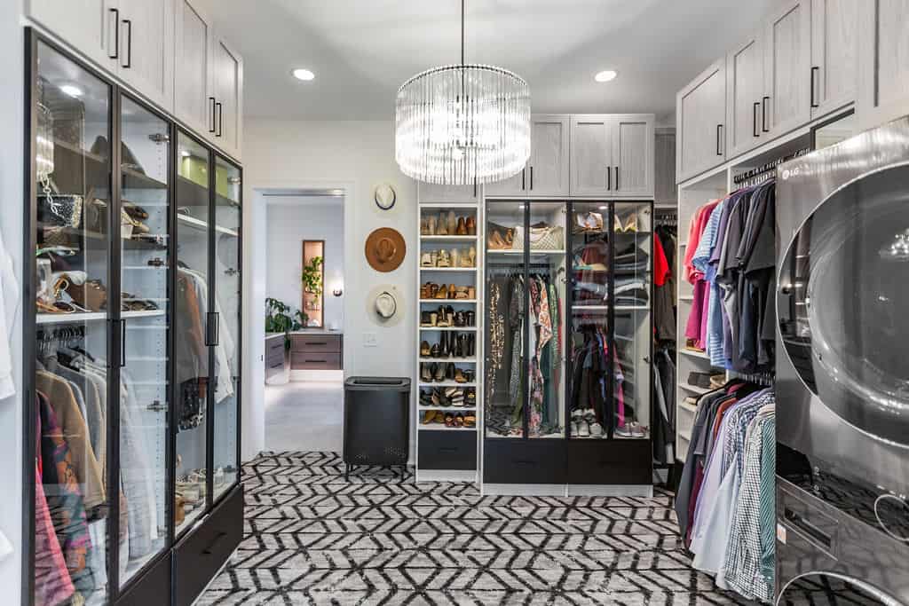 Nicholas Design Build | A well-organized walk-in closet with glass cabinets, shelves holding shoes and accessories, hanging clothes, and a central chandelier. Laundry area with washer and dryer included. Bedroom visible in the background.