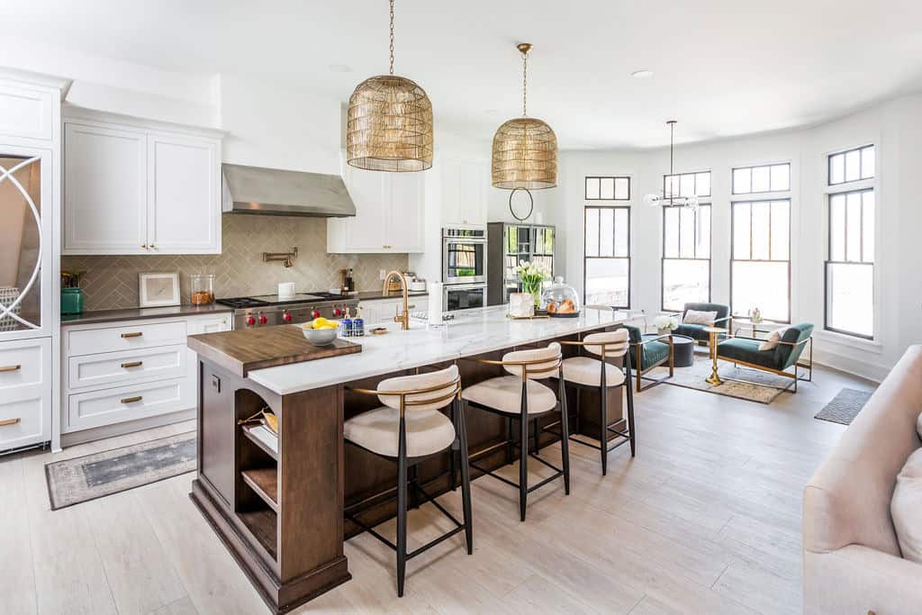 Nicholas Design Build | A modern kitchen with a large island and bar stools, stainless steel appliances, white cabinetry, pendant lighting, and a nearby living area with large windows and contemporary furniture.