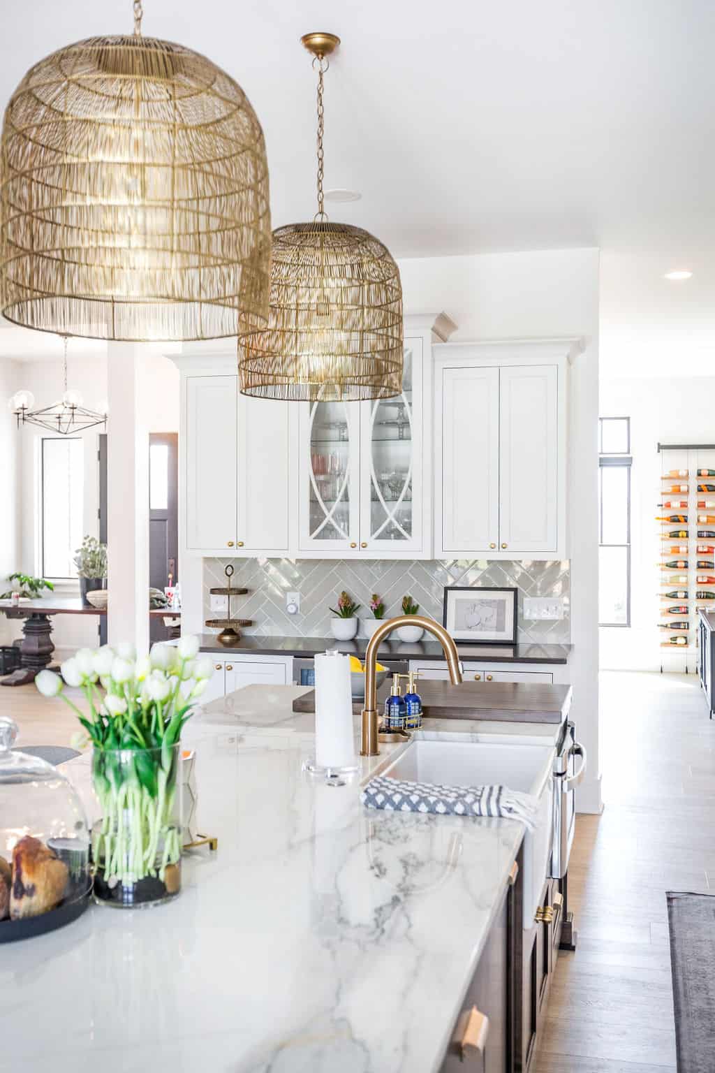 Nicholas Design Build | A modern kitchen with large wicker pendant lights, white cabinets, marble countertops, and a decorative backsplash. A vase of white tulips and pastries are on the island, with a wine rack visible in the background.