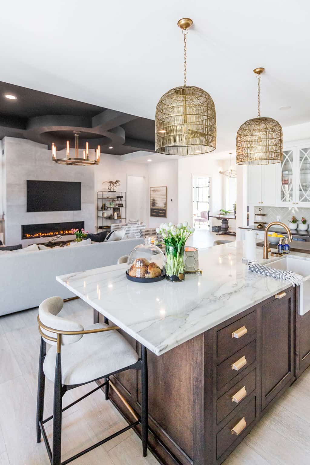 Nicholas Design Build | Modern open-concept kitchen and living area with marble island, gold fixtures, pendant lights, and a large sofa in front of a flat-screen TV and fireplace.