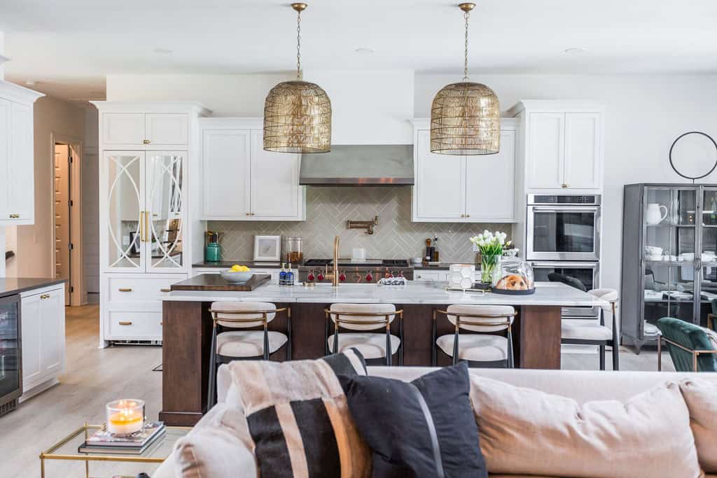 Nicholas Design Build | A modern kitchen features a large island with barstools, white cabinetry, stainless steel appliances, and two metallic pendant lights. A cozy living area with a sofa is in the foreground.