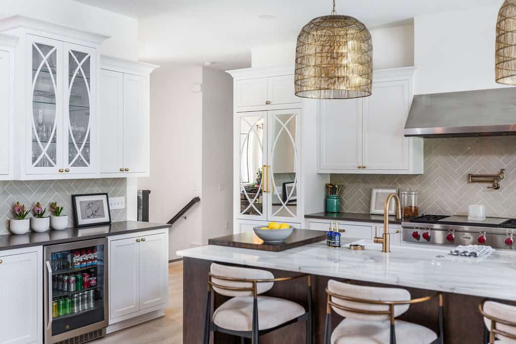 Nicholas Design Build | Modern kitchen with white cabinetry, a marble island with stools, stainless steel appliances, and geometric pendant lights. The background reveals stairs and a mix of open shelving and closed cabinets.