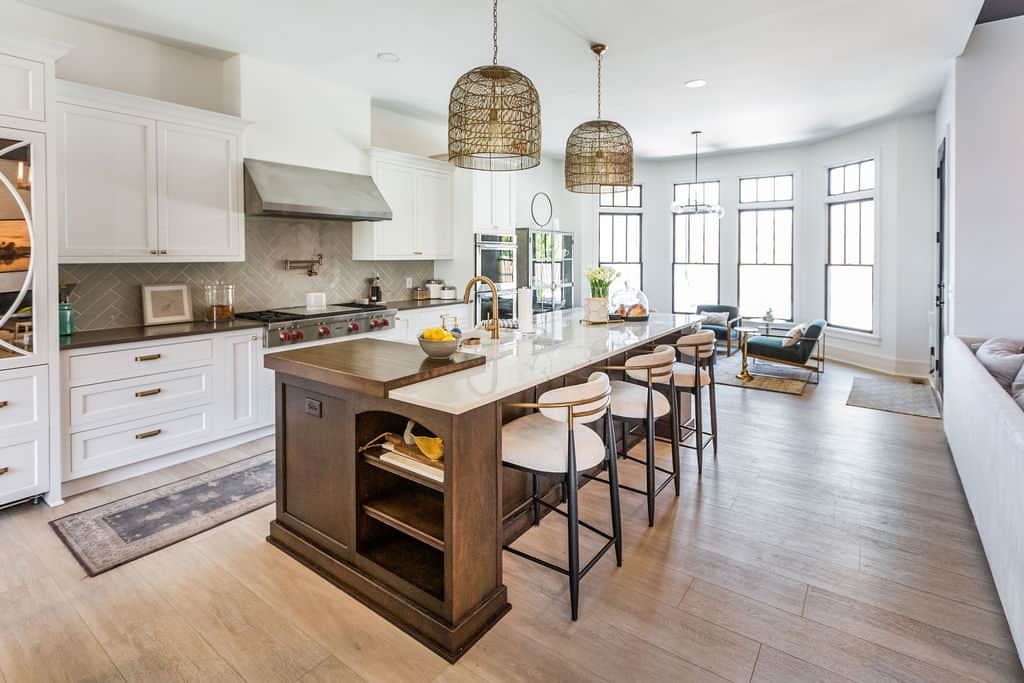 Nicholas Design Build | Modern kitchen with white cabinetry, stainless steel appliances, and a large island with seating for four. Decorative pendant lights hang above the island, and a living area is visible in the background.