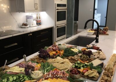 Nicholas Design Build | A kitchen island features an elaborate charcuterie board with various vegetables, fruits, cheeses, crackers, and meats. The background shows a modern kitchen with an oven, sink, and cabinets.