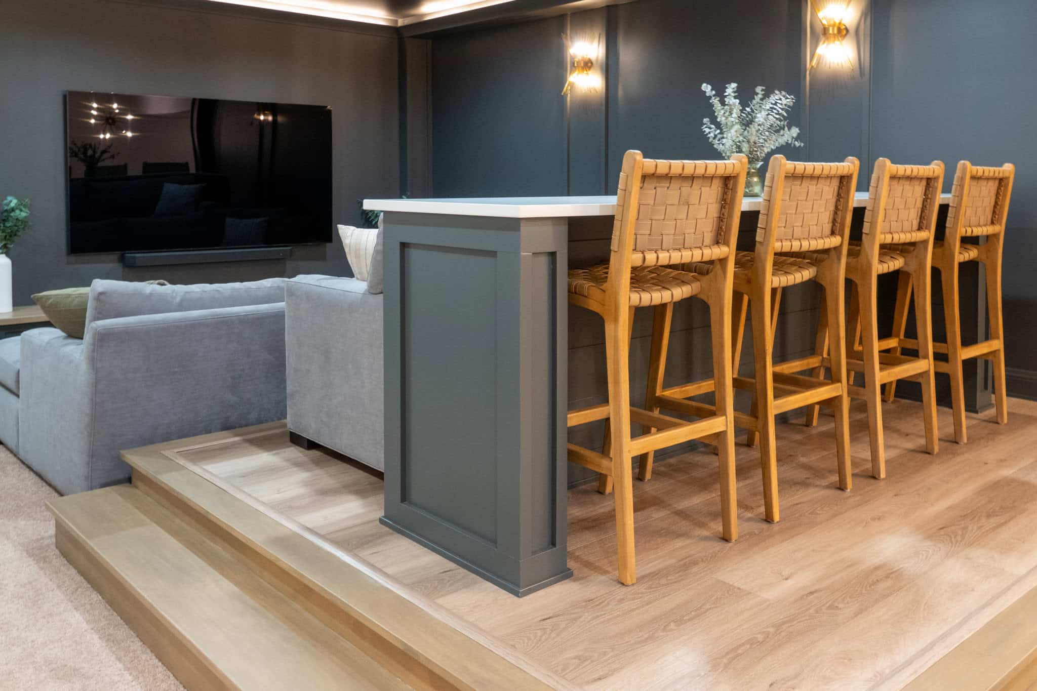 Nicholas Design Build | A modern kitchen and living area with a raised platform featuring a bar counter with wicker chairs, a large TV on the wall, a gray sectional sofa, and minimalist decor.