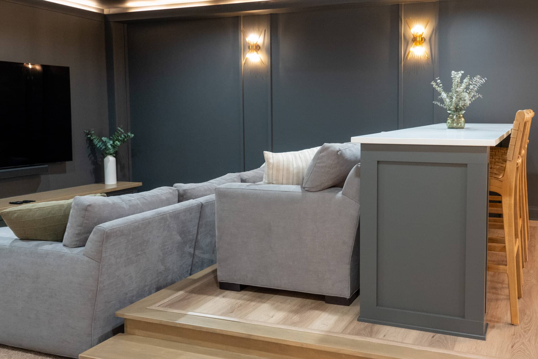 Nicholas Design Build | A modern living room with a grey sectional couch, light grey armchair, wooden coffee table, and a raised bar area with high chairs. The wall has two sconces and a large TV is mounted on the left.