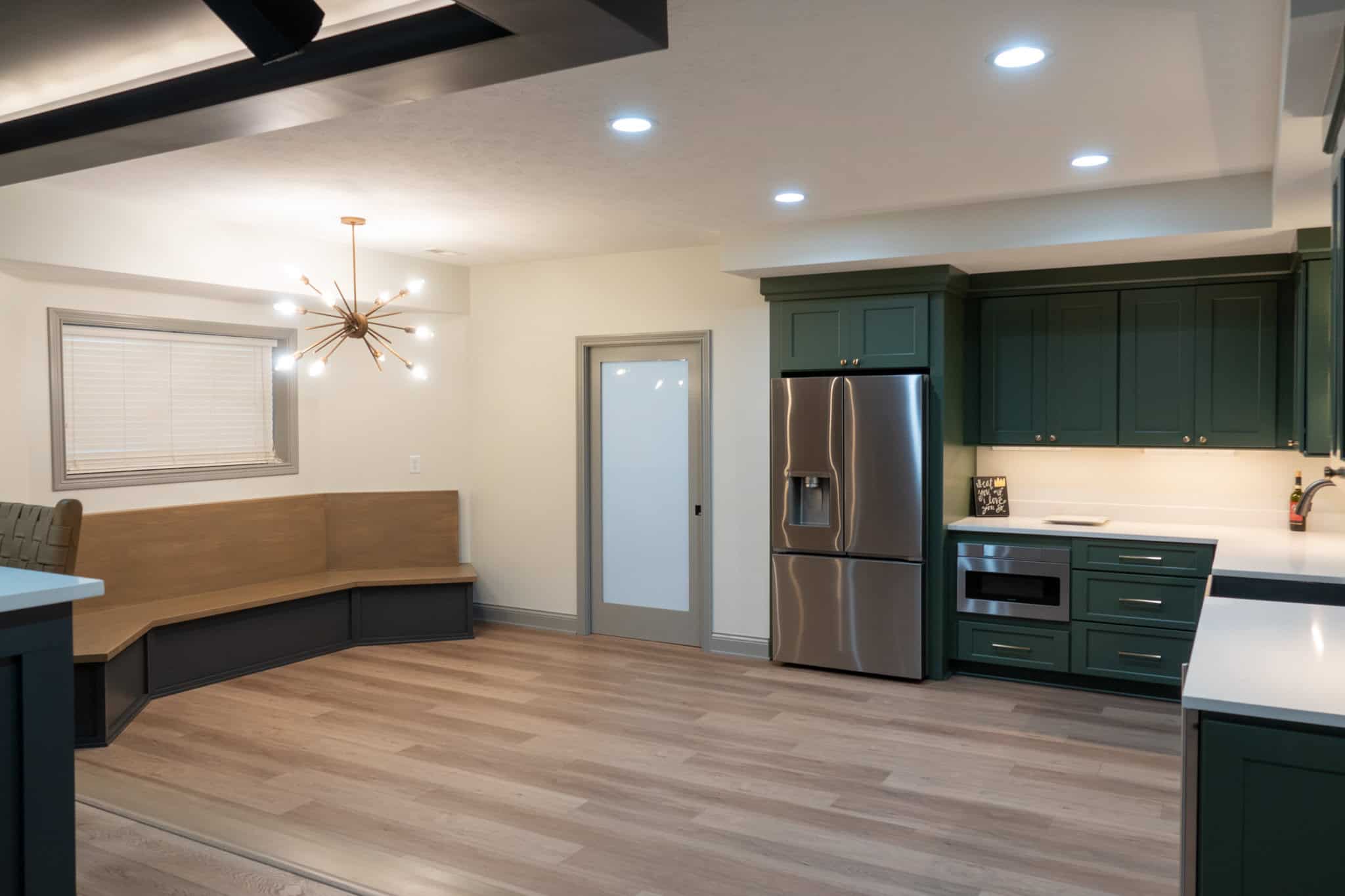 Nicholas Design Build | Modern kitchen with green cabinets, stainless steel appliances, and light wood flooring. A built-in bench with a light fixture is in the dining area to the left.