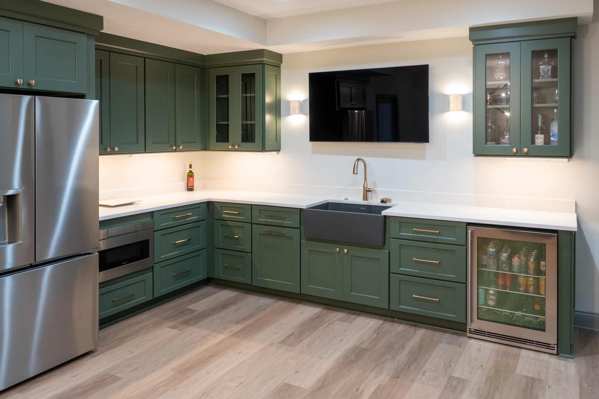 Nicholas Design Build | A modern kitchen with green cabinets, white countertops, stainless steel appliances, a large sink, a beverage cooler, and a wall-mounted TV. The flooring is wood.