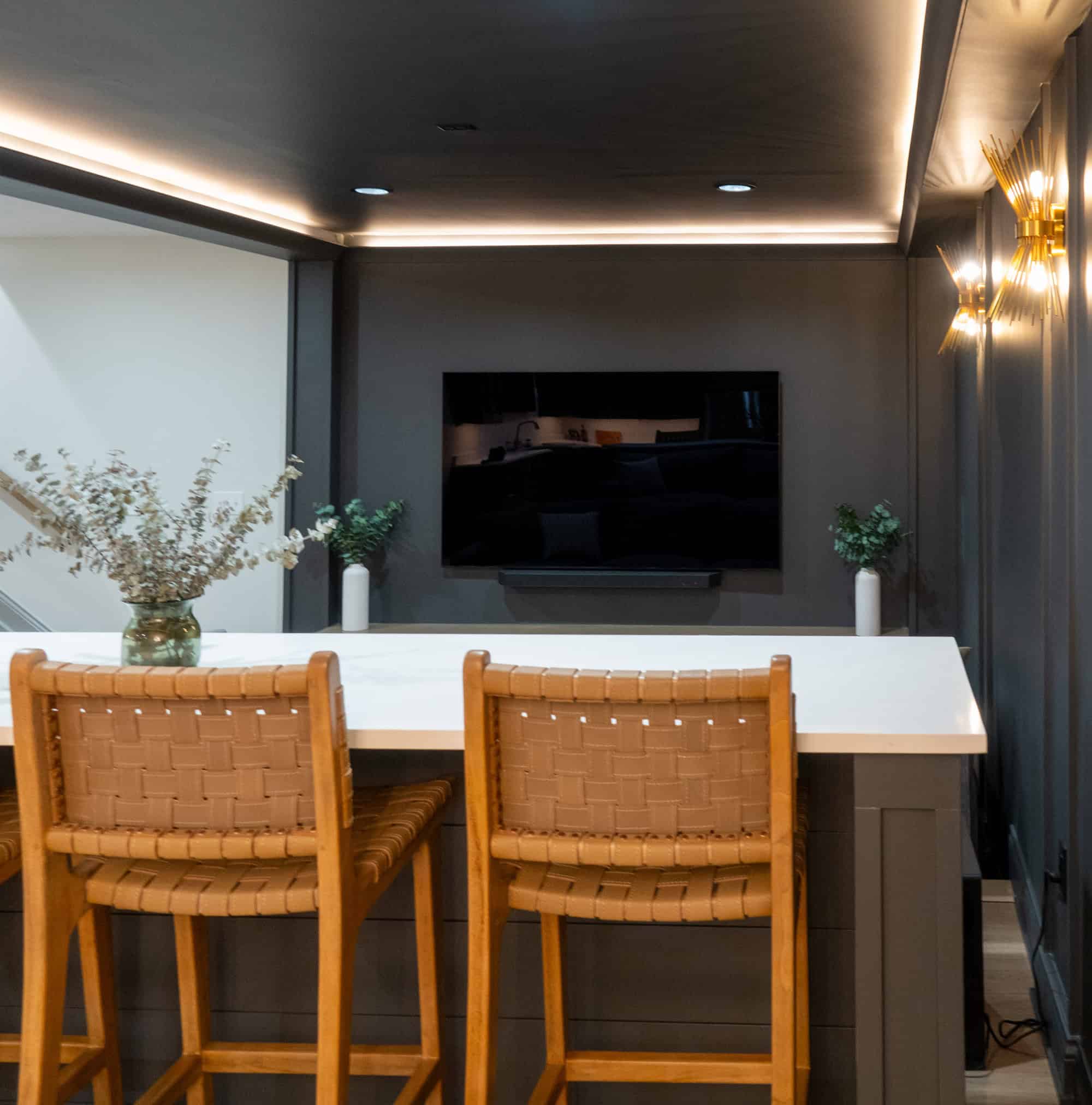 Nicholas Design Build | A modern kitchen area with a white countertop, two woven-back wooden barstools, a mounted flat-screen TV, and decorative plants on the counter and near the TV.