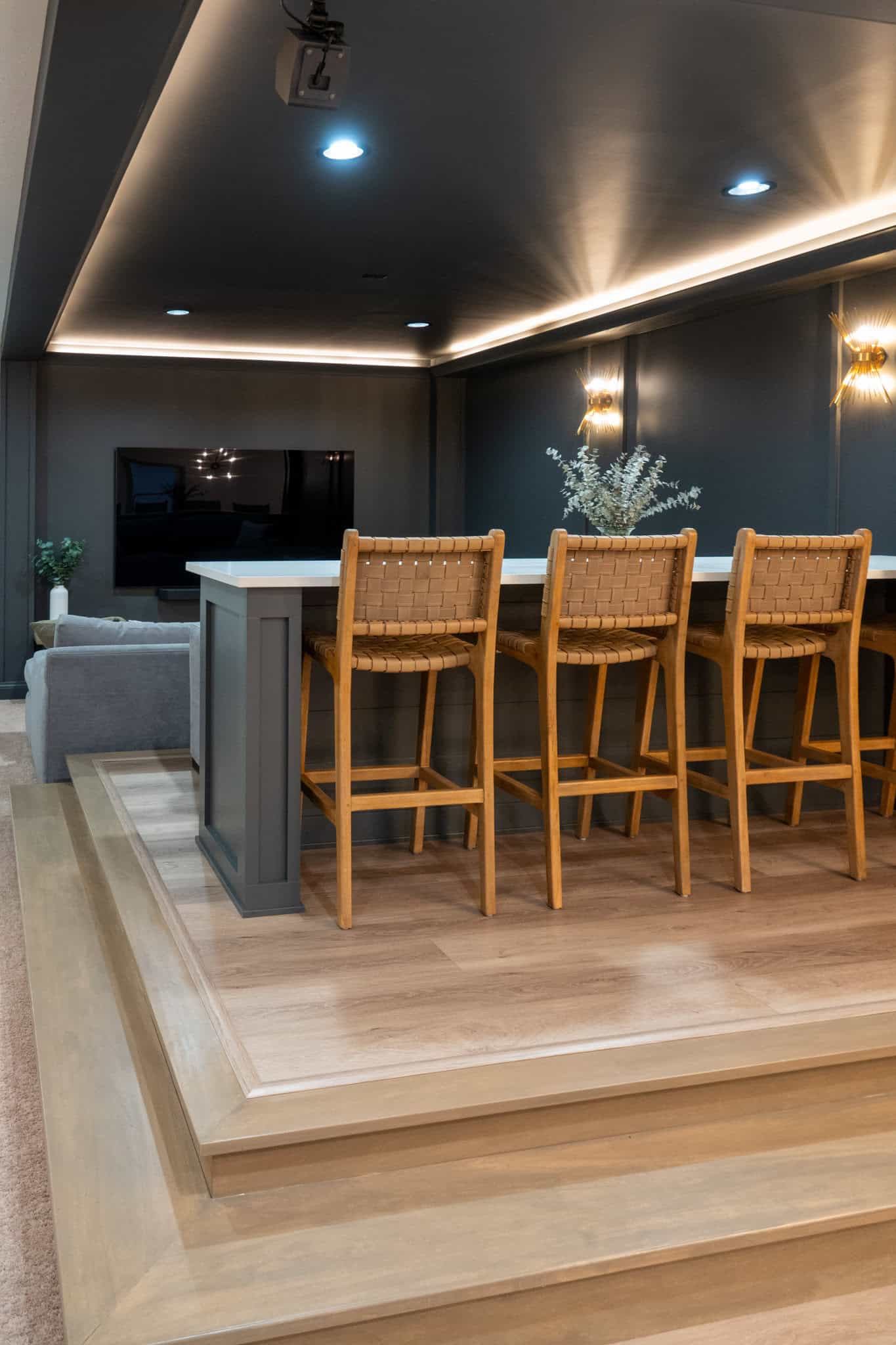 Nicholas Design Build | A home theater room with a large screen, a row of high wooden chairs at a bar, and a gray couch on a raised wooden platform. The room has dim lighting and decor includes wall sconces and potted plants.