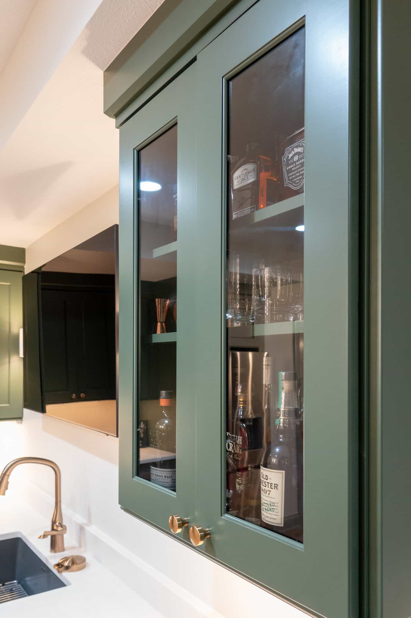 Nicholas Design Build | A kitchen with green cabinets, some featuring glass doors, revealing liquor bottles and glasses inside. A white countertop with a silver faucet is in the foreground.