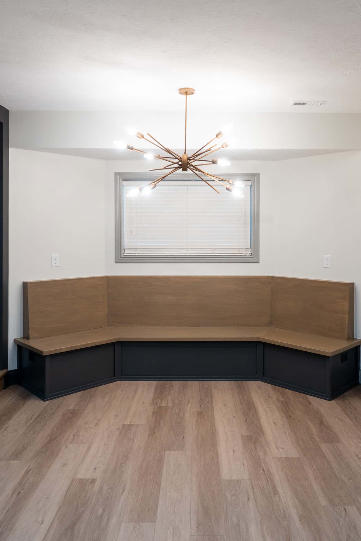 Nicholas Design Build | A minimalist room with a modern chandelier, a corner wooden bench seating, and wood flooring. A single window with blinds is centered above the bench.