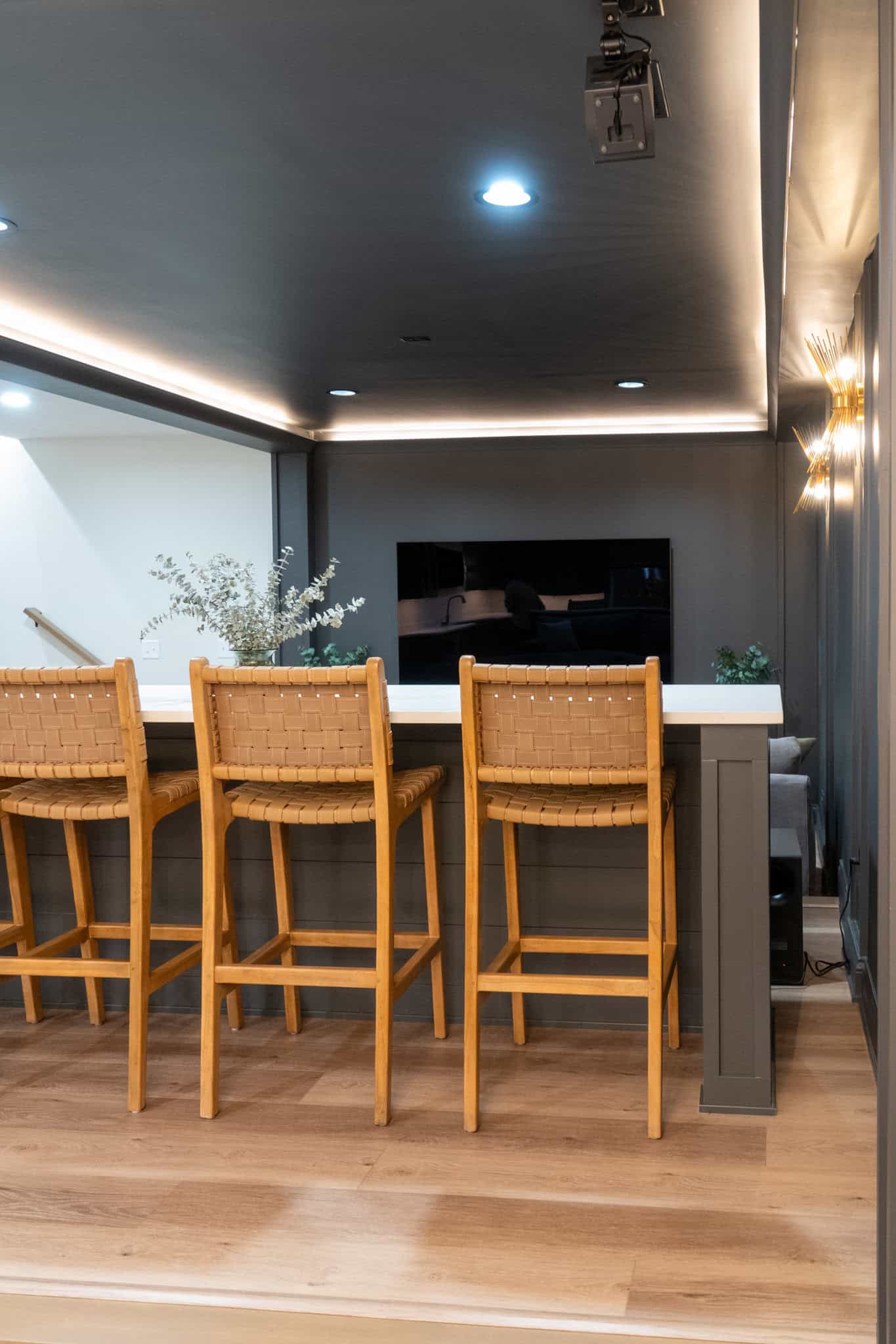 Nicholas Design Build | A modern, dimly lit bar area with three wooden bar stools at a counter. The background features a flat-screen TV, plants, and muted wall lights.