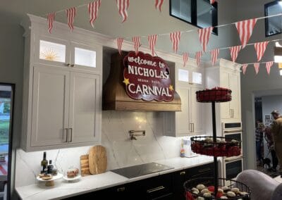 Nicholas Design Build | Kitchen decorated with carnival-themed banners and sign reading “Welcome to Nicholas’ Carnival.” Counters display food and drinks, and cupcakes are arranged on a tiered stand. People are gathered in the background.