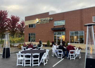 Nicholas Design Build | A group of people sit at white folding tables and chairs outside in front of a brick building with a sign that reads 