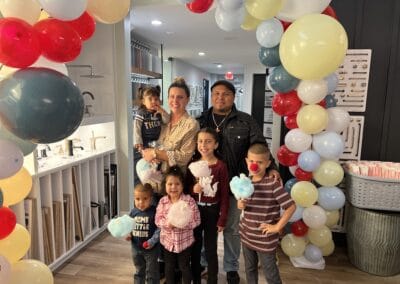 Nicholas Design Build | A family of six stands under a balloon arch. The mother holds a toddler, and the father stands behind three children holding cotton candy. They are indoors, near a counter displaying various items.