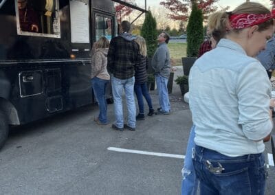 Nicholas Design Build | A group of people standing in line at a black food truck in a parking lot with autumn trees in the background.