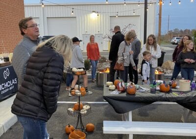 Nicholas Design Build | A group of people, including adults and children, gather outdoors near a table with pumpkins and decorations. Some are looking at the table, and string lights are hung overhead.