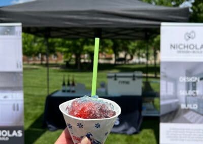 Nicholas Design Build | A hand holds a colorful snow cone in front of an outdoor booth with banners and a tent on a sunny day.