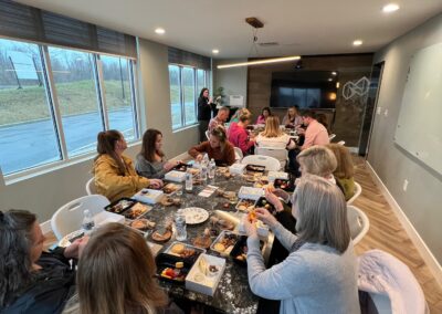 Nicholas Design Build | A group of people are seated at a long table in a conference room, enjoying a meal together. Food containers and drinks are spread out on the table. Large windows and a TV screen are visible in the room.