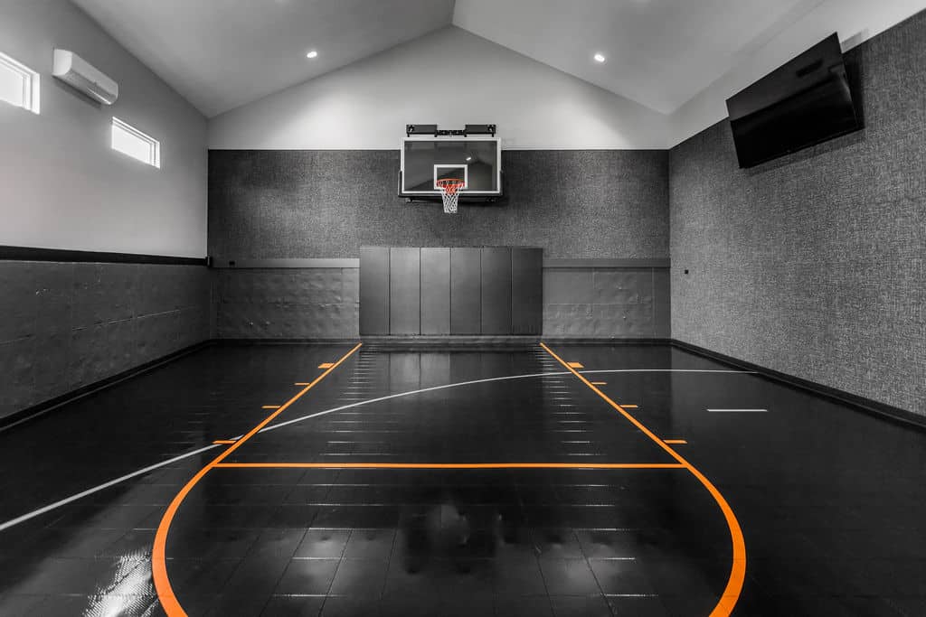 Nicholas Design Build | Indoor basketball court with a hoop, black flooring, and orange lines. Gray walls and a large TV screen are visible. Ceiling lights illuminate the space.