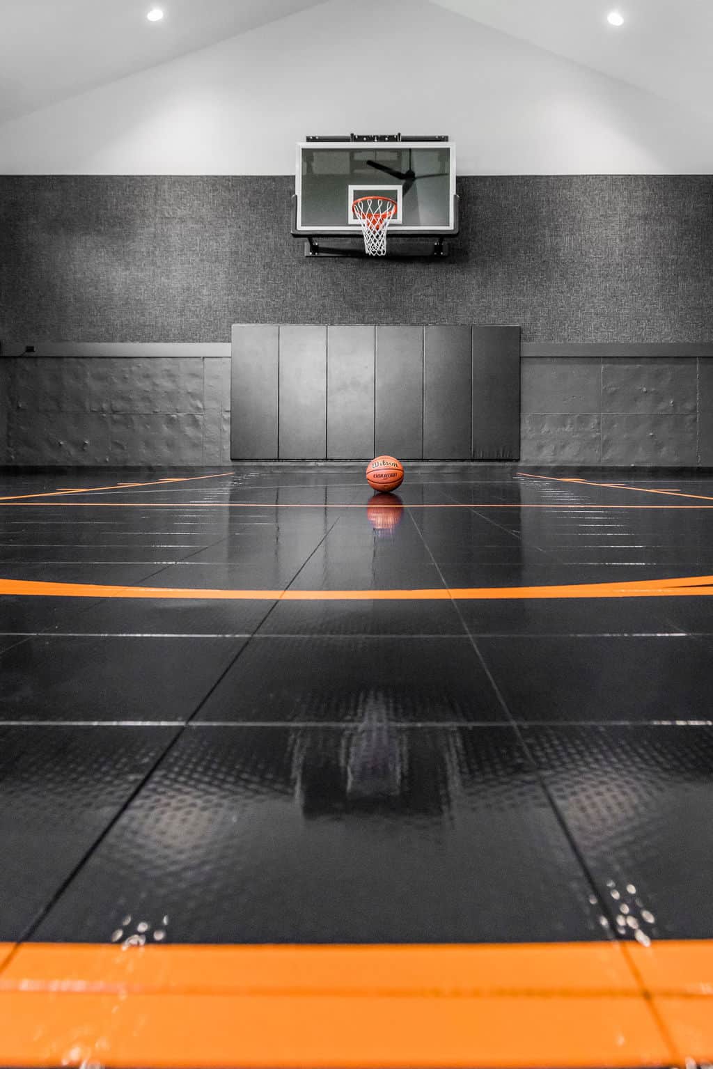 Nicholas Design Build | A basketball rests at the center of an indoor court with an orange and black floor, facing a hoop on a wall.