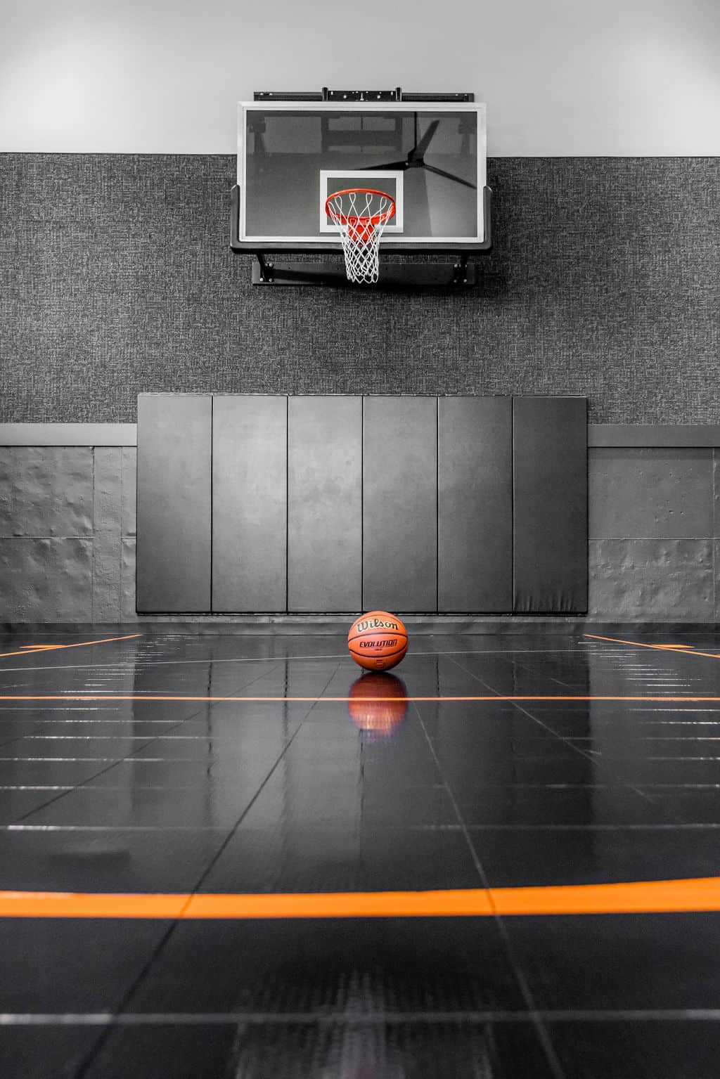 Nicholas Design Build | A basketball positioned on a glossy indoor court in front of a hoop and backboard mounted on a padded wall.