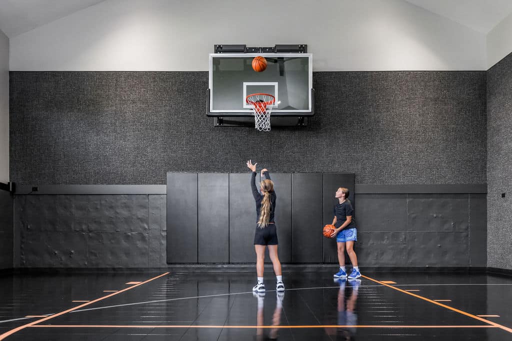 Nicholas Design Build | Two people practicing basketball indoors; one shoots while the other holds a basketball. The court has a dark floor and padded walls.