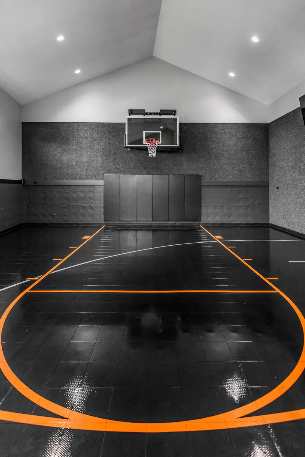 Nicholas Design Build | Indoor basketball court with a black floor and orange lines, featuring a single hoop mounted on a padded wall in a well-lit room.