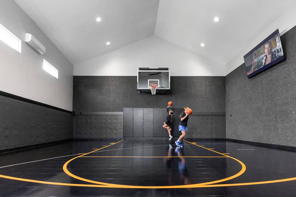 Nicholas Design Build | Two people playing basketball in an indoor court with black and orange flooring, a wall-mounted hoop, and a flat-screen TV.