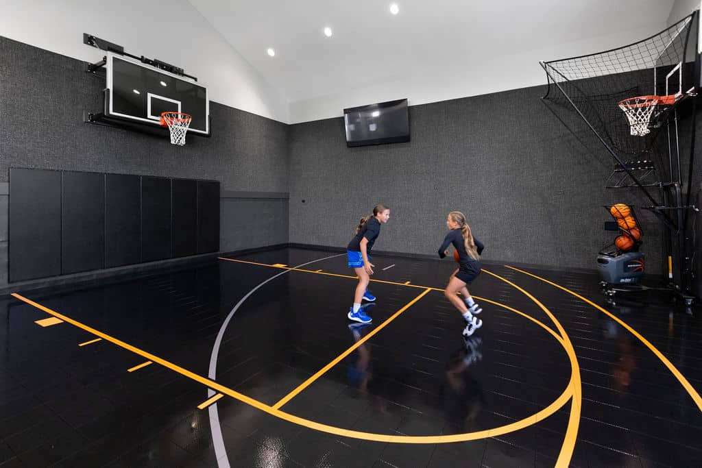 Nicholas Design Build | Two people playing basketball indoors on a black court with a basketball machine and wall-mounted hoops.