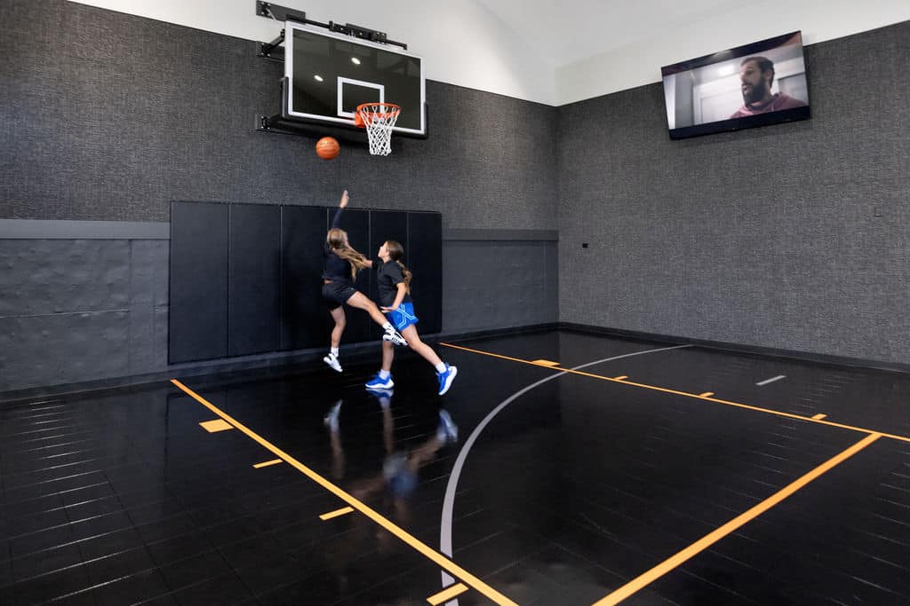 Nicholas Design Build | Two people playing basketball on an indoor court. One is attempting a layup while the other defends. A large screen above shows a person speaking.