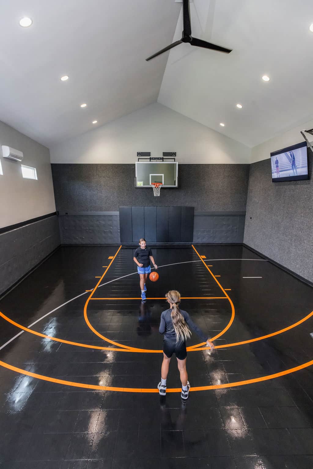 Nicholas Design Build | Two people playing basketball in an indoor court with black floors and orange lines. A TV is mounted on the wall.