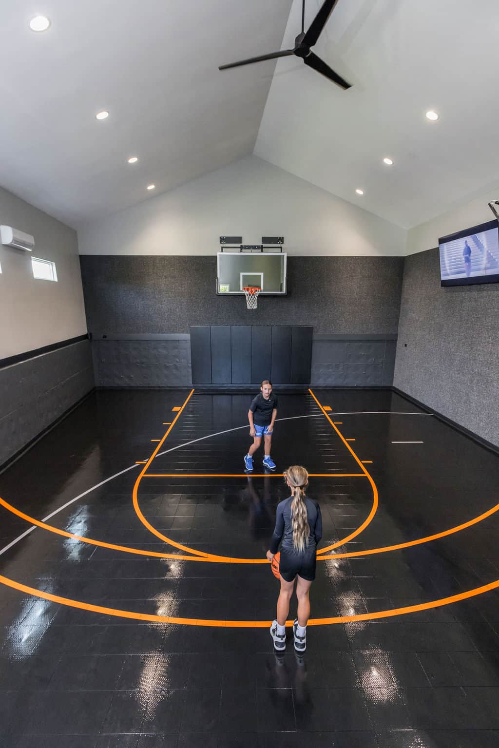 Nicholas Design Build | Two people playing basketball in an indoor court with a glossy black floor and orange lines, under bright overhead lights and a ceiling fan.