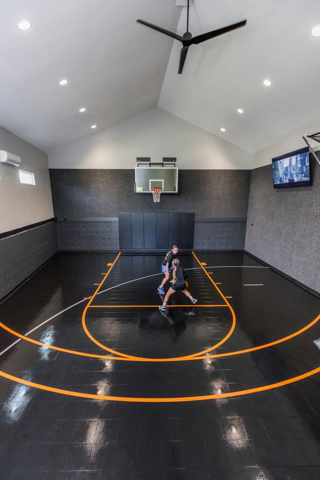 Nicholas Design Build | Two people playing basketball on an indoor court, with a black polished floor and orange lines, under bright ceiling lights and a ceiling fan. A television is mounted on the wall.