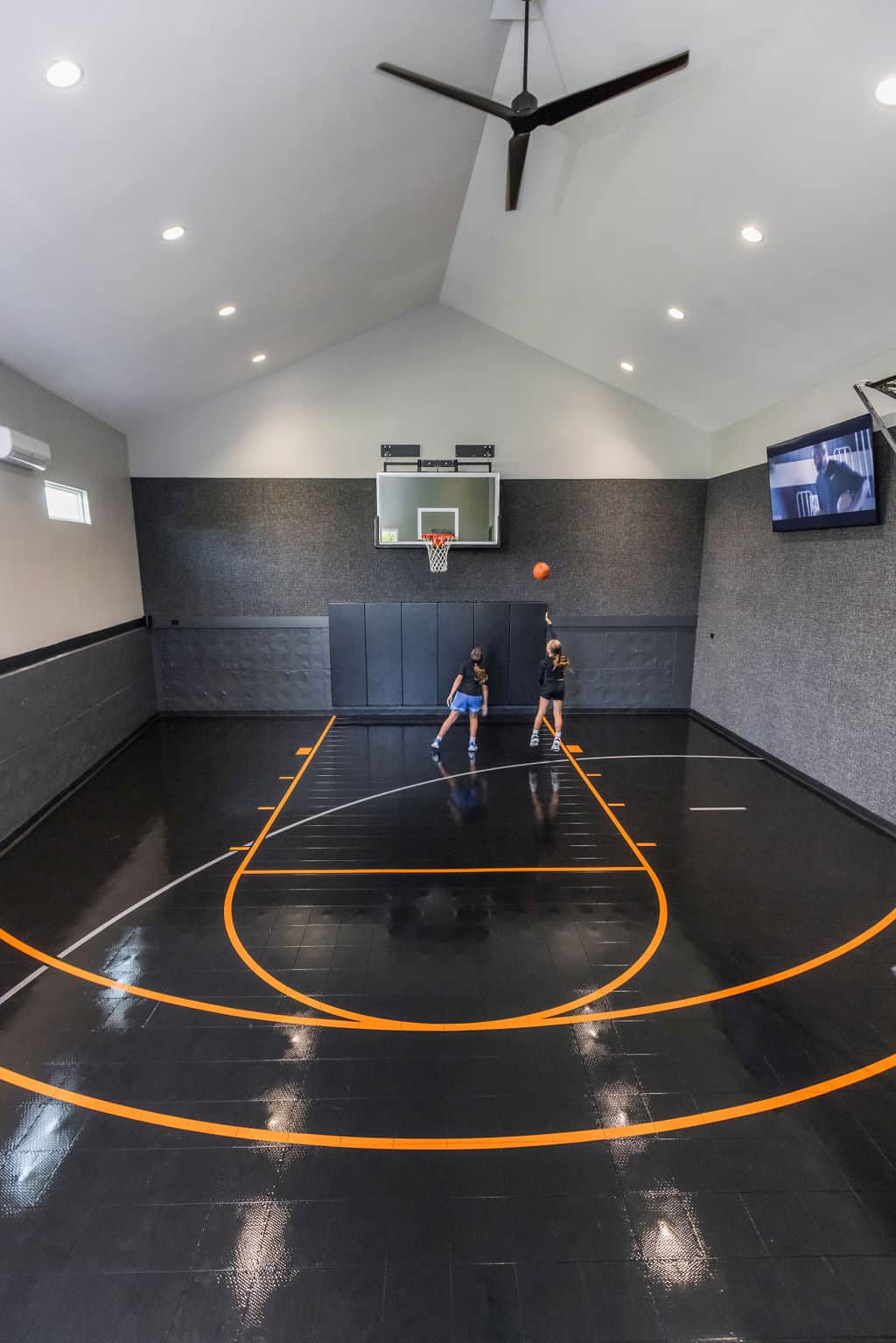 Nicholas Design Build | Two people play basketball on a polished indoor court with black and orange lines. A wall-mounted hoop, ceiling fan, and flat-screen TV are visible.