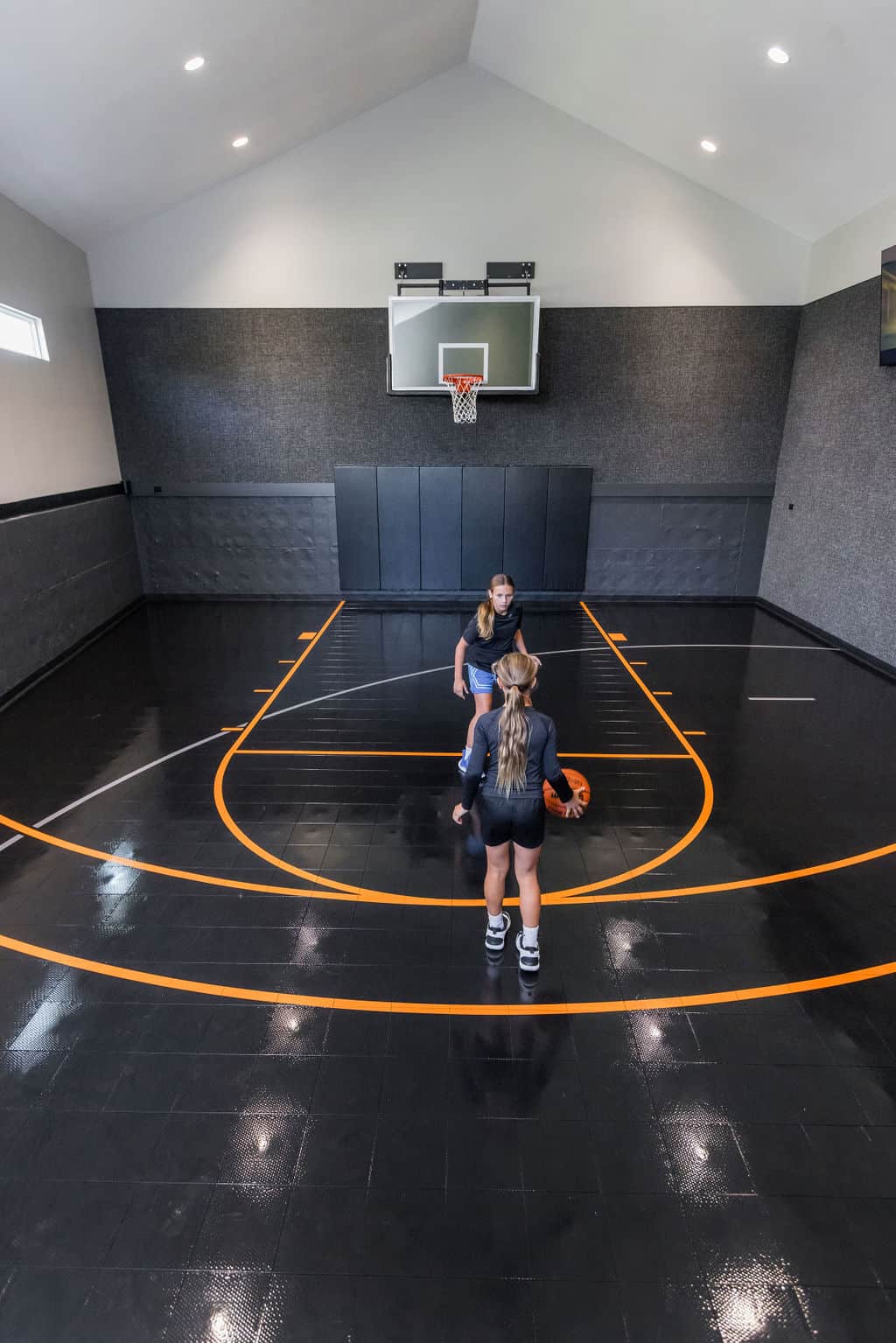 Nicholas Design Build | Two people playing basketball indoors on a black court with orange lines, near a basket.